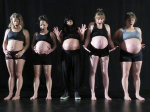 Five pregnant women standing in a line, looking down at their bumps in suprise, as if they have only just noticed they are pregnant.