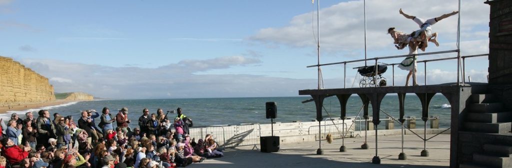 A large outdoor audience looking onto a raised stage with 3 acrobats performing on it with a baby crib on the stage with them. In the background is the English Channel on a sunny day.