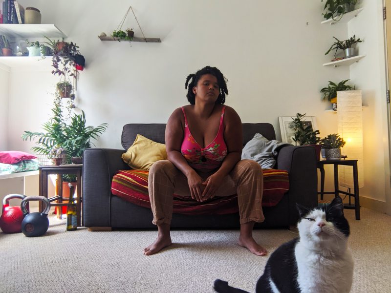 A mixed race woman in her early twenties sitting on her sofa and looks straight in the camera, she wears beige trousers and a bright pink bikini top, She has a cool and unimpressed facial expression with her elbows resting on her knees. In the background are a variety of potted plants, kettlebells, books and glass bottles. A white and black spotted cat sits looking into the camera in the right hand corner.