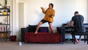 A mixed race woman her early thirties, in an ochre loose knit jumper and black shorts dancing on her black couch and a burgundy throw rug couch.