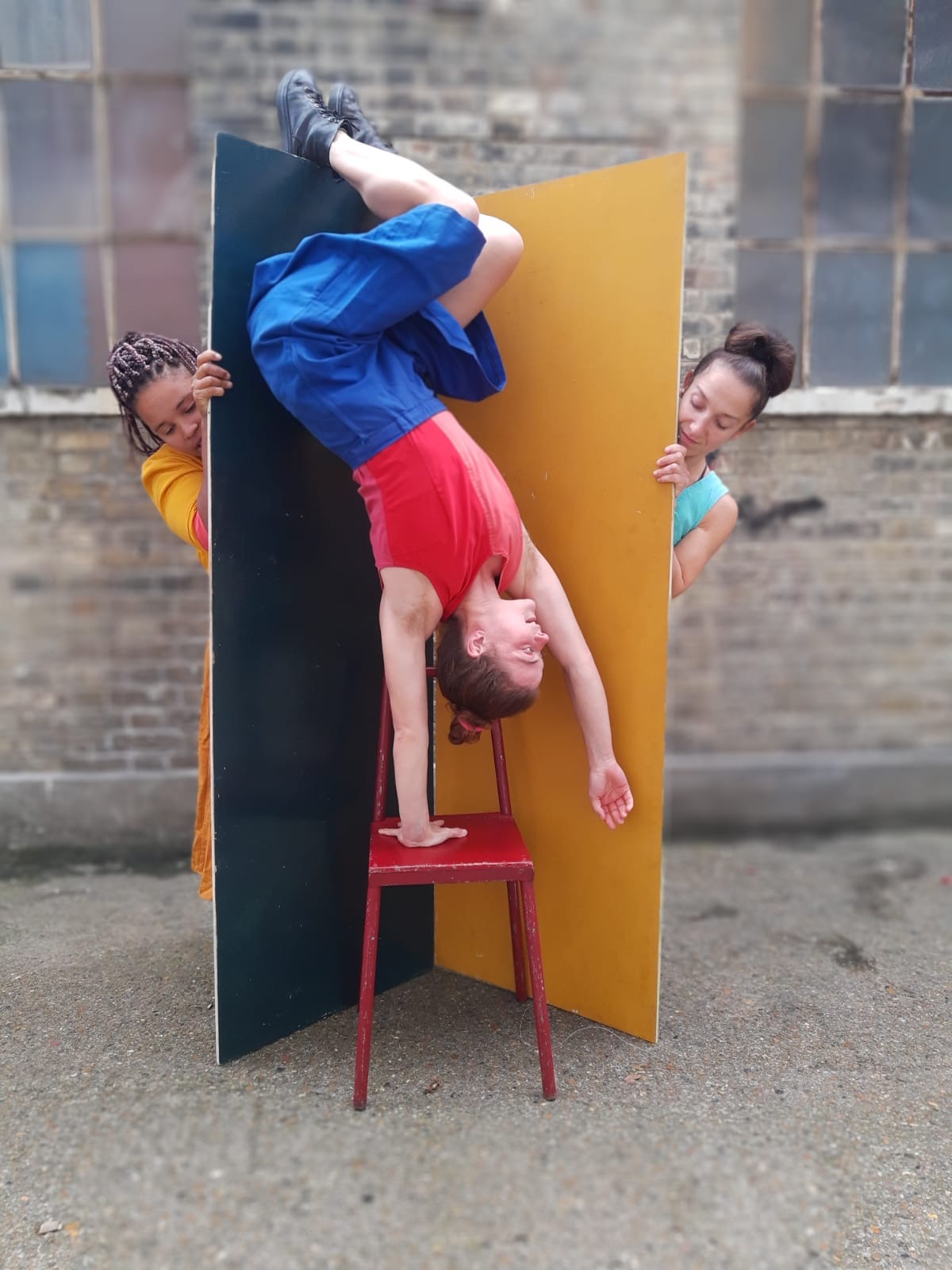 a woman doing a handstand on a chair with two women l=behind her holding up wide planks and looking at the first woman from behind the prop walls.