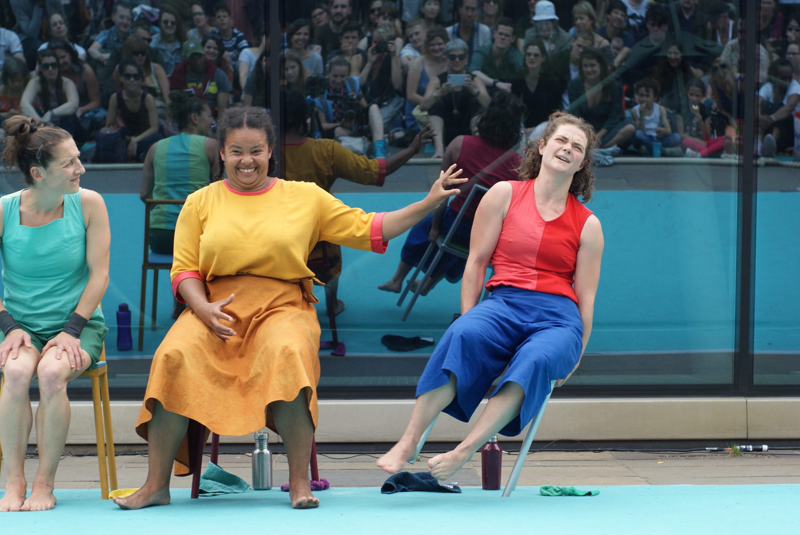 three women sitting on chairs. One of them is toppling to the side after being pushed by the woman sitting next to her. In the background is a reflects a large outdoor audience.