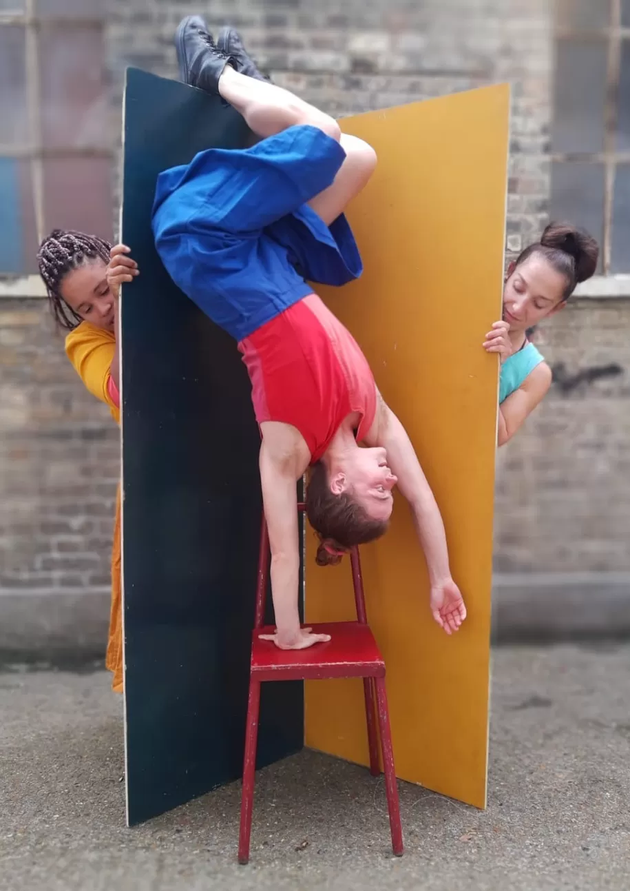 a woman doing a handstand on a chair with two women l=behind her holding up wide planks and looking at the first woman from behind the prop walls.