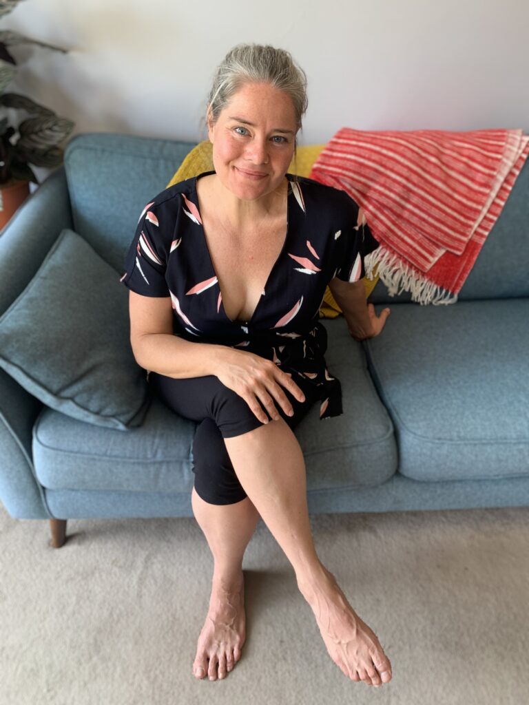A middle-aged woman sits on a duck egg blue sofa wearing black leggings and a black top with white and light plink patters. She looks into the camera with a warm smile. Behind her a crimson and white patterned throw rug and a mustard yellow throw rug rest on the back of the sofa.