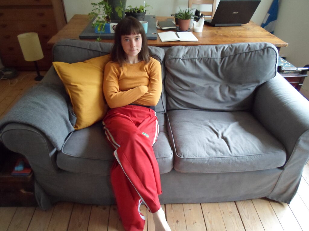 a young woman sits on her light grey sofa looking into the camera, she wears an ochre long sleeve top and red trousers with white stripes along the outside seams of the legs. Behind her is a desk with notebooks, an open laptop and plants. 