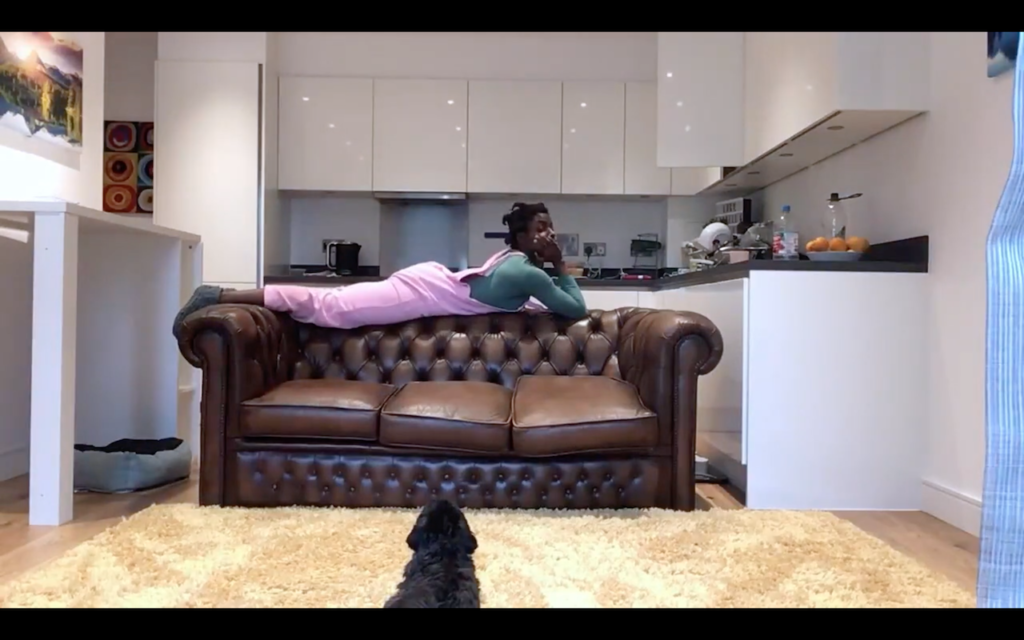a young woman lies on her front on the back of her brown leather sofa with her head propped up on her hands. She wears light pink dungarees, a teal long sleeves top and thick grey socks. She sneakily glances down at a small black dog sitting in the foreground of the picture. In the background is the kitchen.