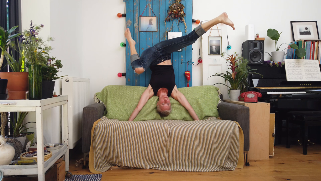 A woman upside down in a hand stand on her couch. She is surrounded by plants and there is a piano to one side.