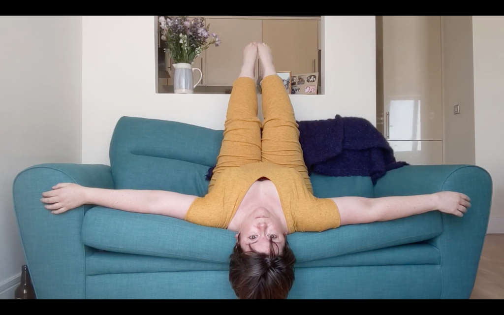 a woman lying upside down in a teal couch wearing a mustard yellow jumpsuit, her legs are raised, and her arms rest agains the front of the armrests