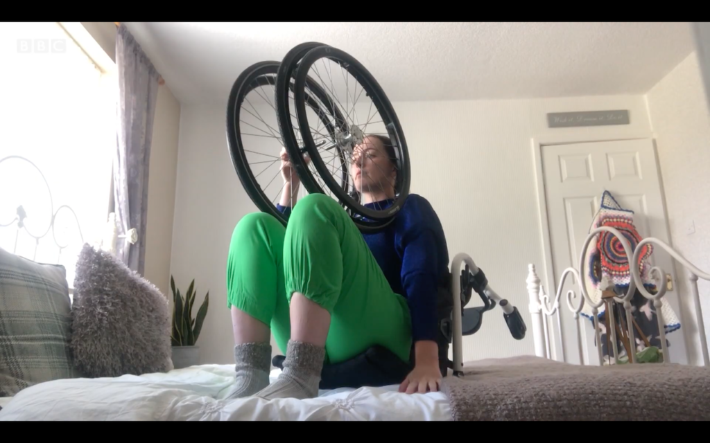 A woman siting on her bed on the backrest of her dissamebled wheelchair. She holds the wheels on either side of her face. She wears bright green trousers a blue sweater and thick grey socks.
