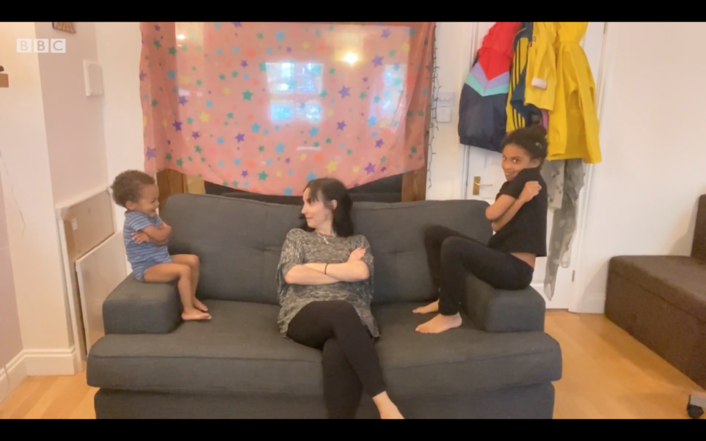 A woman sitting in a grey couch with her young teen daughter on one end of the couch, sitting on the armrest and the woman's toddler on the other end of the couch. All three of them have their arms crossed, the children are smiling.