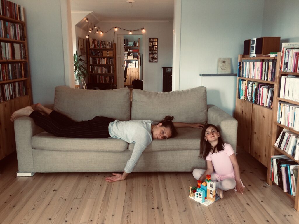 a woman lies on her stomach on her sandstone couch looking tired with her hand trailing down on the floor. Her daughter in a light pink ballerina dress sits on the floor with a lego house in front of her. Around them are bookshelves filled with books and CDs.