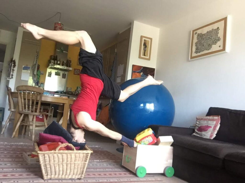 A woman in a headstand with her head in a basket and her arms straight in front of her holding a caddy box with children's toys in it behind her is a kitchette.