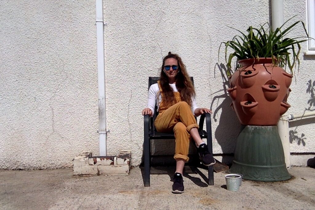 A woman sitting on a black sunchair with her arms on the armrest. She is wearing sunglasses mustard coloured dungarees and a whit long sleeve top underneath. She is outside against a white wall with a large terracotta plant pot with a plant in it.