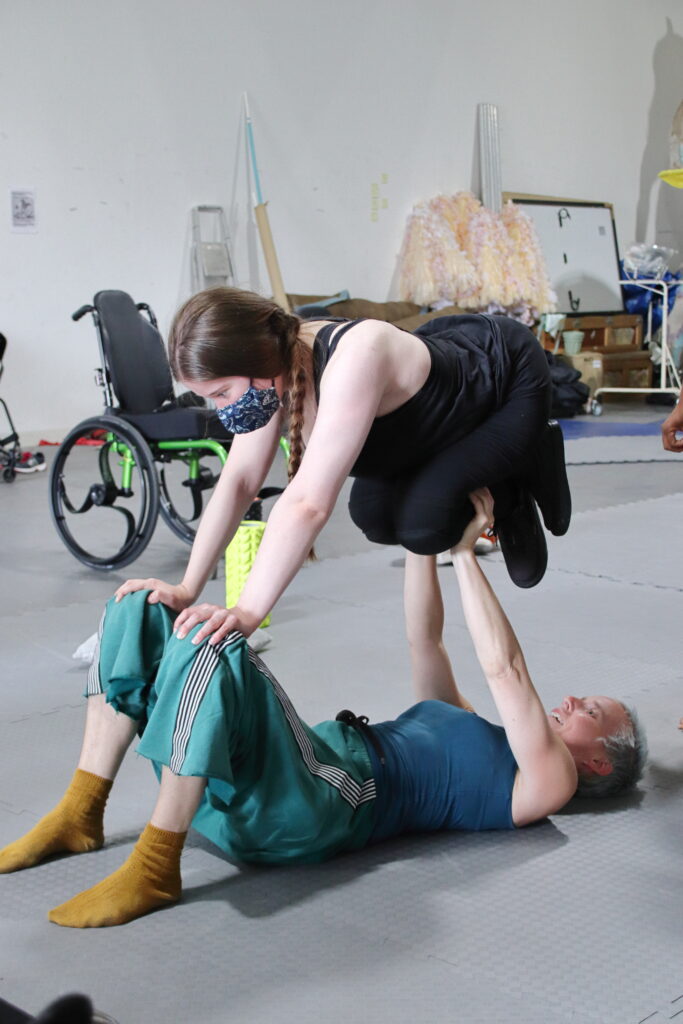 Scene from the Inclusive Acrobalance Reisdancy. Two women perform and acrobalance trick. One of them lies on her back with knees propped up, lifting the other woman's knees. The other woman , lifted in the air supports her hands on the first woman's knees. There are props grey and blue mats in the background and a wheelchair with an electric green frame.