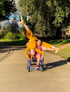 A wheelchair using woman holds the ankles of a white woman, who is doing a handstand holding onto the base of the wheelchair with her legs in a split. Theyy are in a bright sunny park.