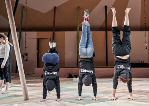 3 women doing a handstand. the one on the left has her knees tucked in. All three wear a black hoodie with white writing on the back "Mimbre Acrobat". They a indoors on a pastel, dusty green, cream and peach checkered floor.