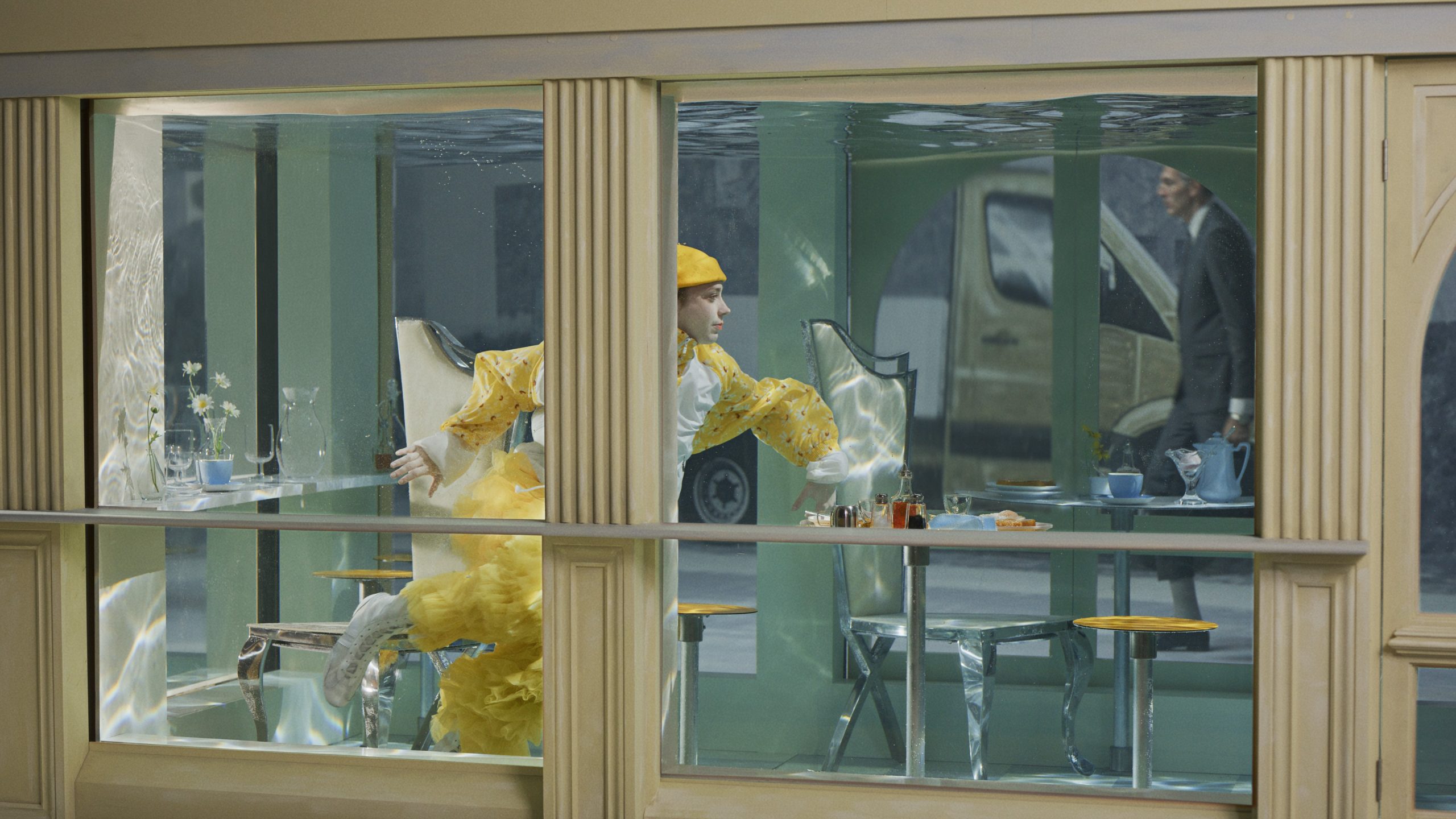 A still from The Glass Hermit. It shows a performer wearing yellow moving through a cafe filled with water.