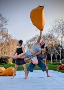 Three female acrobats perform in a park. Two are lifting another who lies across them lifting a big orange bag over her head. They wear grey and are performing on a grey carpet.