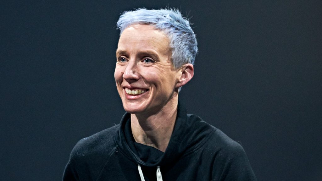 A white woman with hair dyed light blue smiles, looking slightly to the right. She is wearing a black hoodie and is against a dark background.