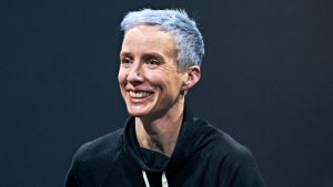 A white woman with hair dyed light blue smiles, looking slightly to the right. She is wearing a black hoodie and is against a dark background.