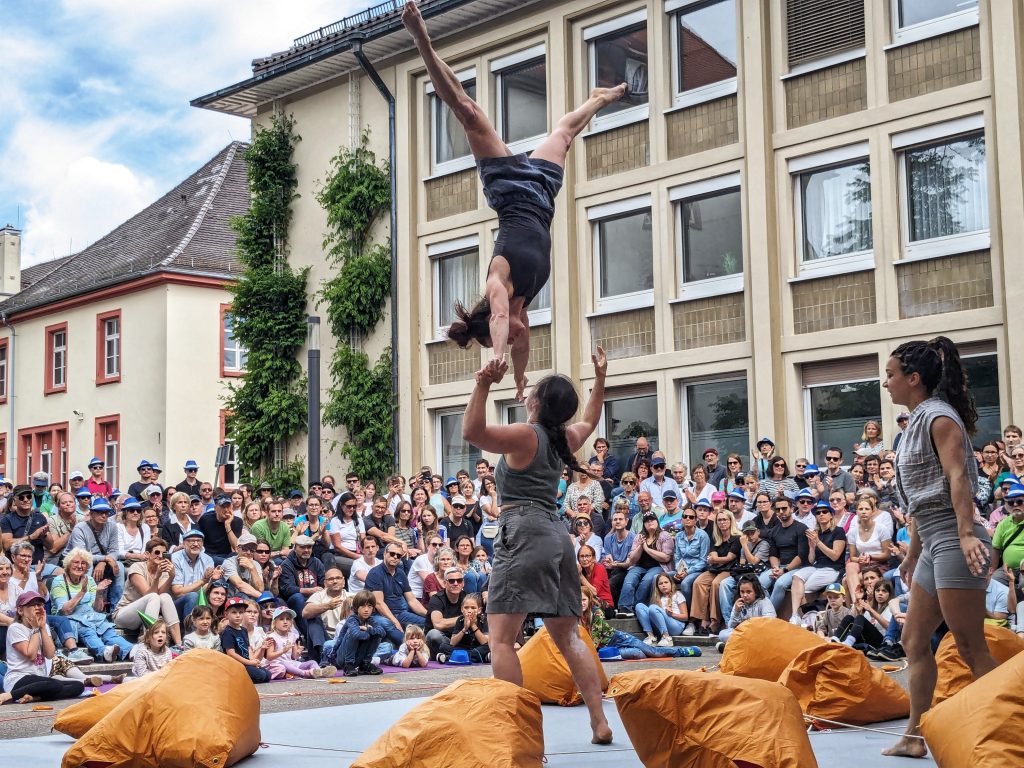 An acrobat wearing grey shorts and a top being thrown up in the air by another acrobat. They are both in front of a big crowd. Orange bags are dotted around the floor around them.