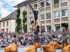 An acrobat wearing grey shorts and a top being thrown up in the air by another acrobat. They are both in front of a big crowd. Orange bags are dotted around the floor around them.