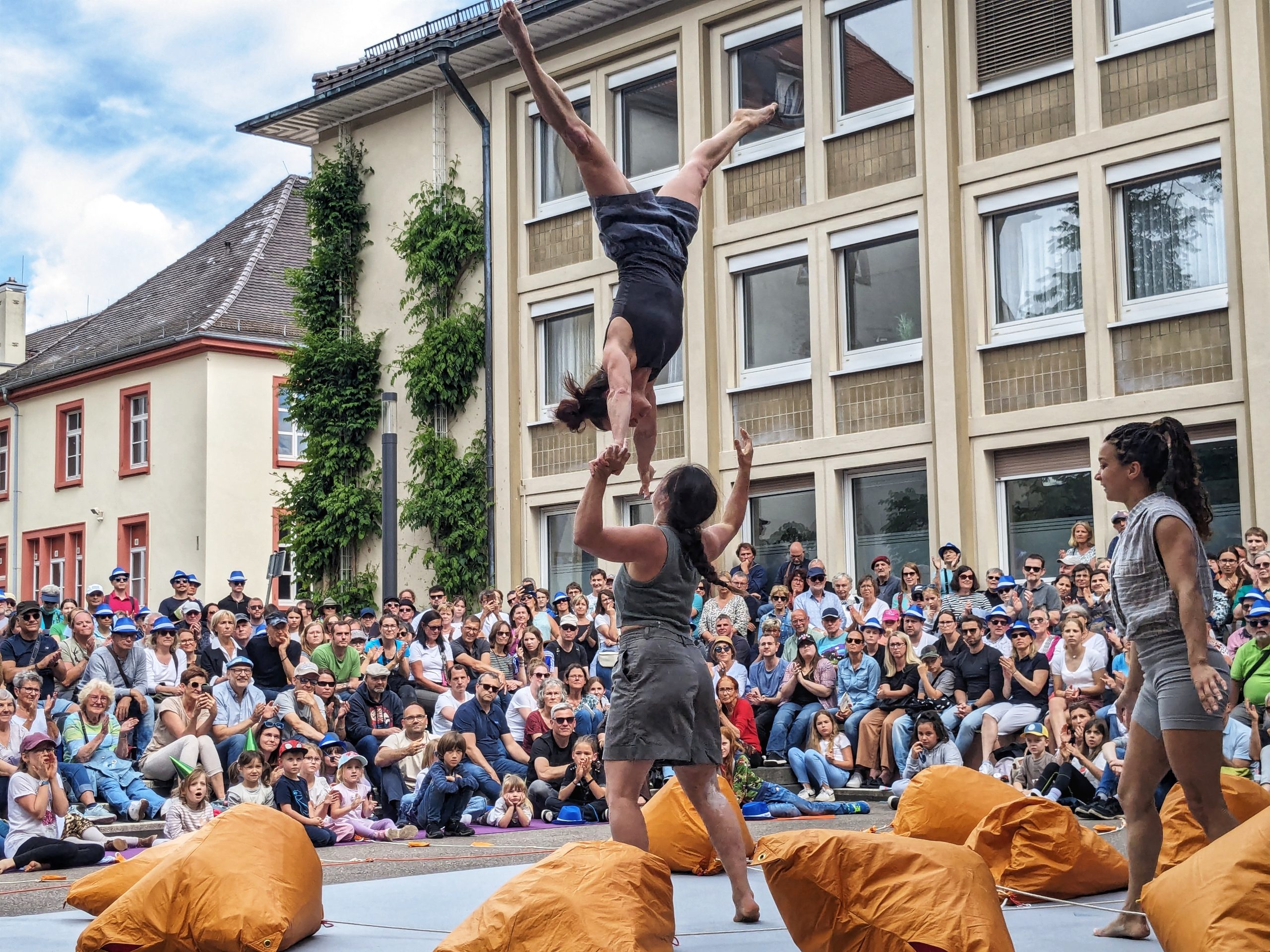 An acrobat wearing grey shorts and a top being thrown up in the air by another acrobat. They are both in front of a big crowd. Orange bags are dotted around the floor around them.