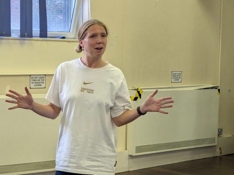 A picture of a young white woman in a community hall. She is explaining something with her arms wide.