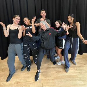 Five dance students and their tutor posing for the camera and looking like they’re in a band. They all wear dark blue, black or grey clothing, are in front of a black curtain, and stand on a wooden floor.