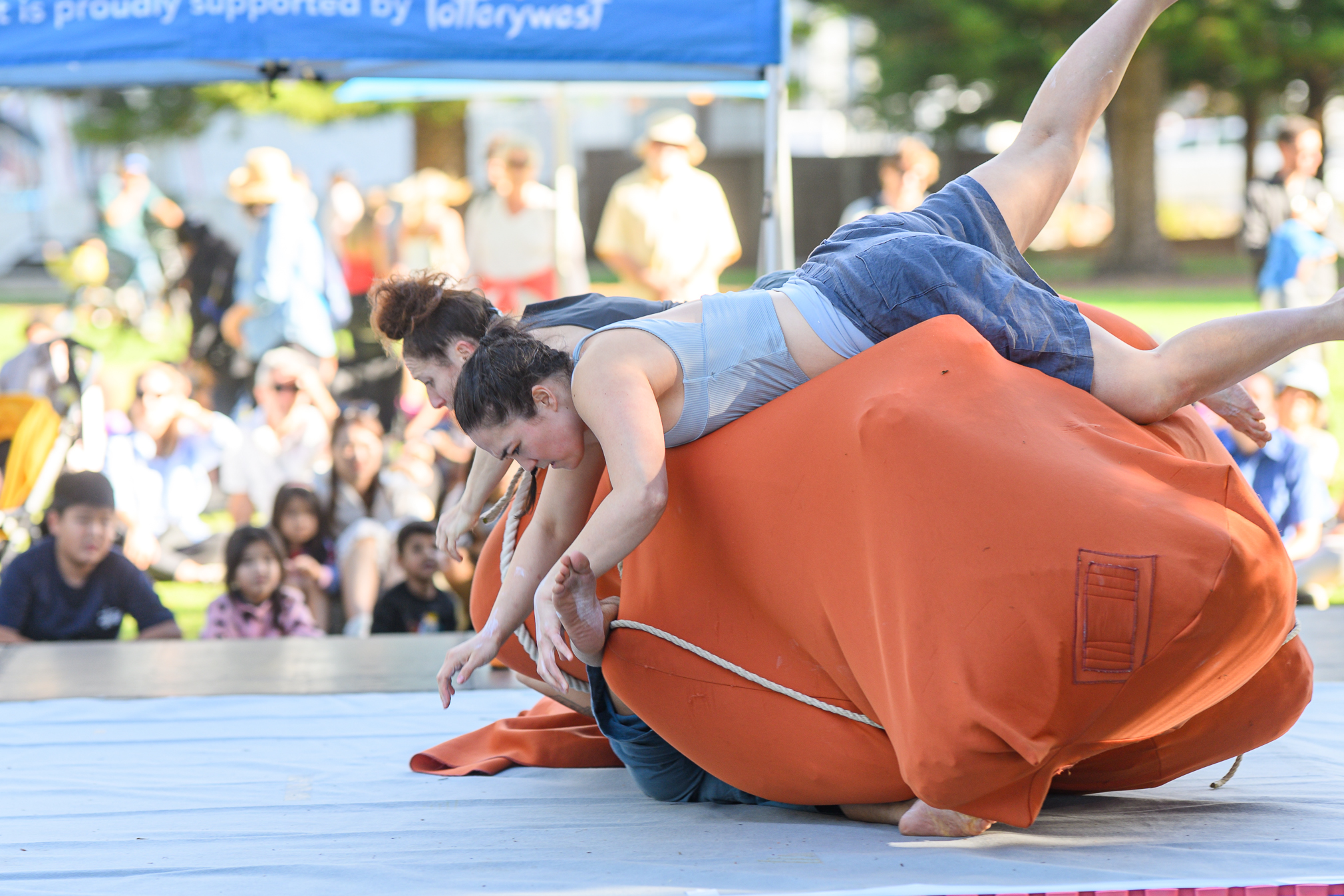 Two women dressed in grey tops and shorts are rolling forwards over a big orange bag. Behind them an audience sitting on some sunny grass can just be seen.