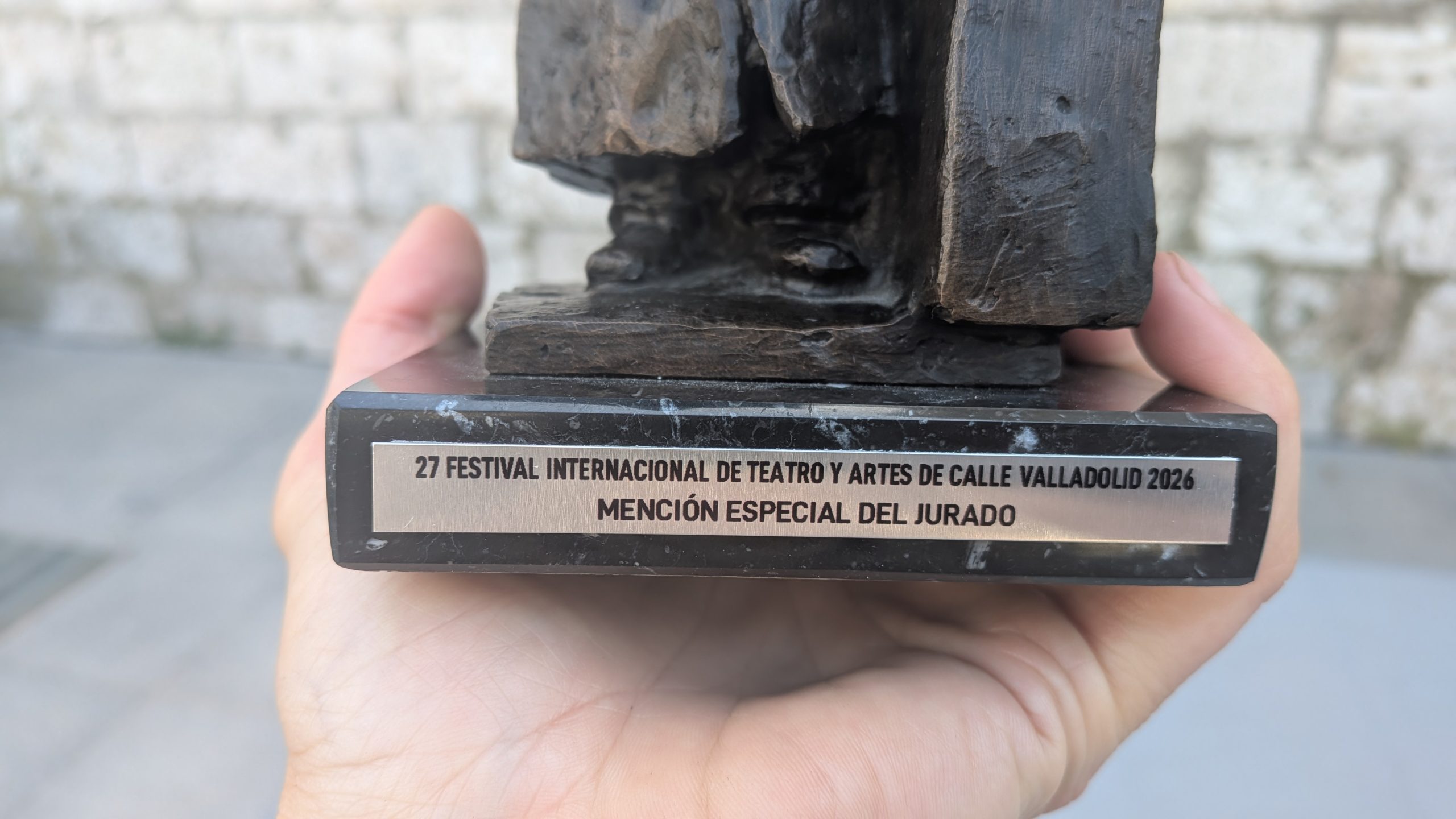 A picture of the bottom of an award. It says: "27 Festival Internacional De Teatro Y Artes De Calle Valladolid 2026. Mención Especial Del Jurado". We can also see the hand holding the award.