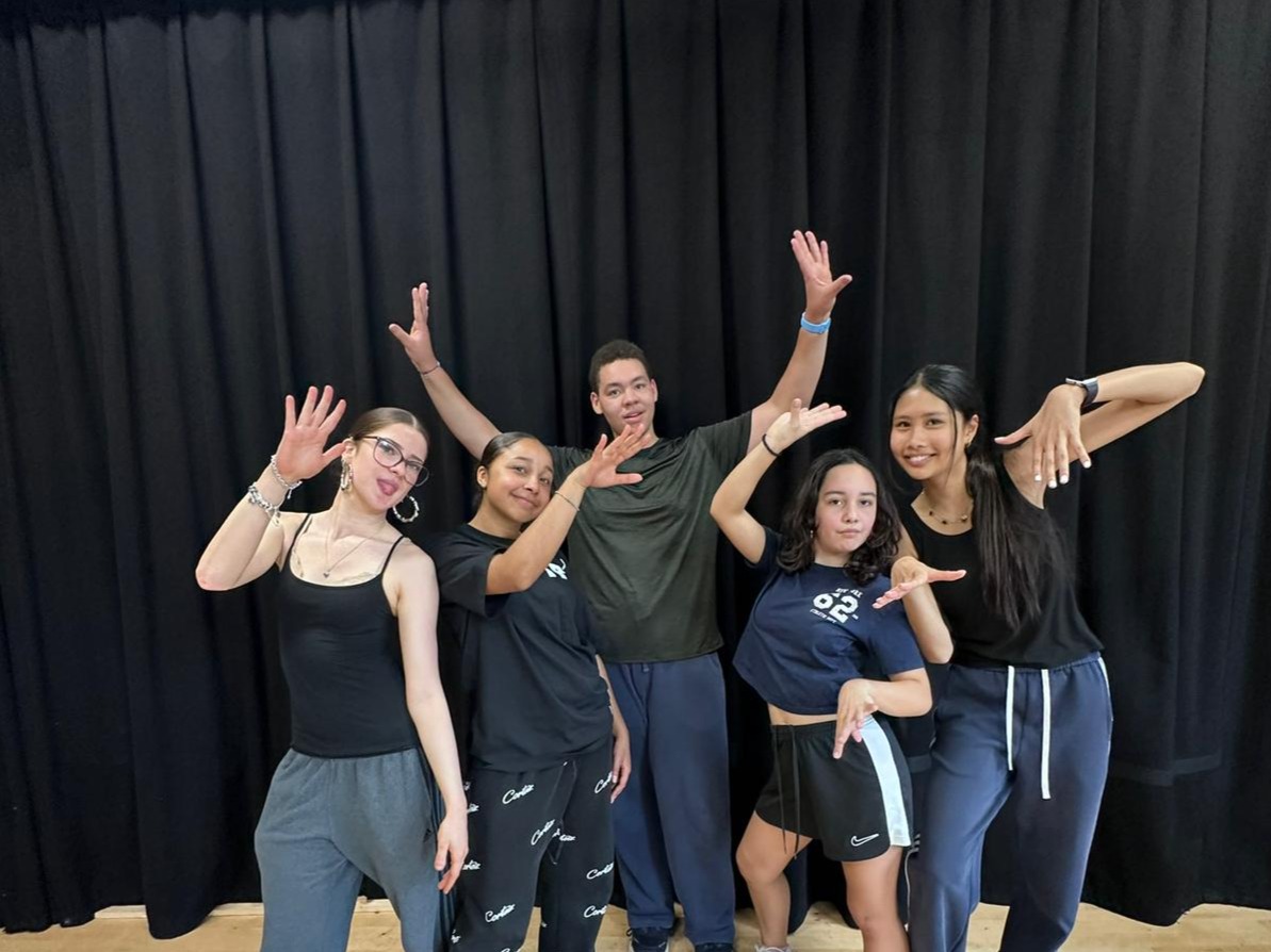 Five dance students posing for the camera and looking like they’re in a band. They all wear dark blue, black or grey clothing and are in front of a black curtain.